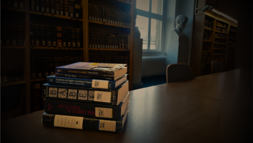 A stack of dark academia-themed books, being the 6 titles introduced and linked in the text, lie on a wooden desk in the Historical Building of Göttingen State and University Library. The mood is dark and gloomy. The background shows a window framed by two statues, chairs and rows of old books on wooden shelves